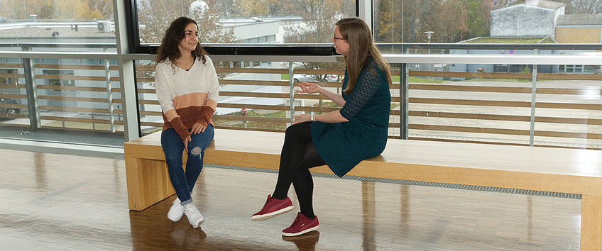 Two doctoral students sit on a bench in the institute and talk to each other.